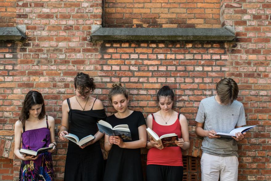 vijf jongeren lezen een boek, ze staan naast elkaar en leunen tegen een bakstenen muur